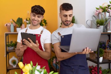 Two hispanic men florists using laptop and touchpad working at flower shop