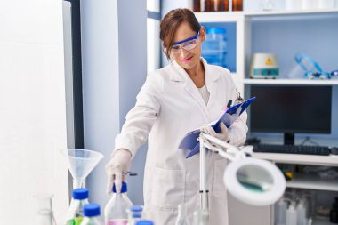 Middle age woman wearing scientist uniform holding clipboard at laboratory