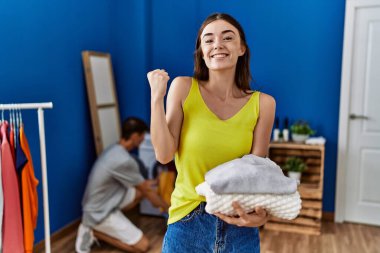 Young brunette woman holding folded laundry screaming proud, celebrating victory and success very excited with raised arms 