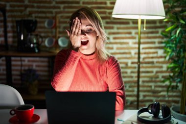 Blonde woman using laptop at night at home covering one eye with hand, confident smile on face and surprise emotion. 