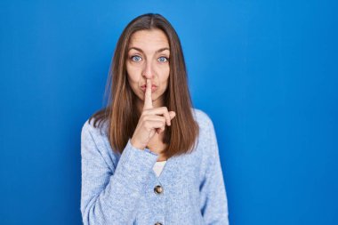 Mavi arka planda dikilen genç bir kadın parmakları dudaklarında sessiz olmak istiyor. sessizlik ve gizli konsept. 