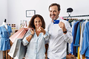 Hispanic middle age couple holding shopping bags and credit card pointing to you and the camera with fingers, smiling positive and cheerful 