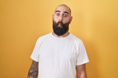Young hispanic man with beard and tattoos standing over yellow background making fish face with lips, crazy and comical gesture. funny expression. 