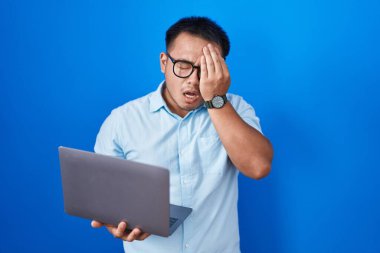 Chinese young man using computer laptop yawning tired covering half face, eye and mouth with hand. face hurts in pain. 