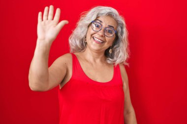 Middle age woman with grey hair standing over red background waiving saying hello happy and smiling, friendly welcome gesture 