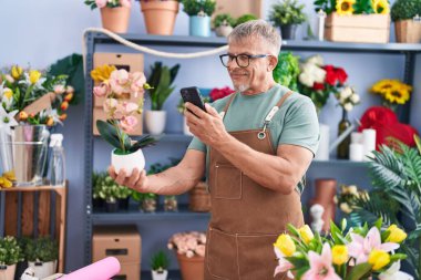 Middle age grey-haired man florist make photo to plant by smartphone at flower shop