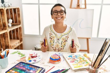 Resim stüdyosunda elinde renkli kalemlerle gülümseyen İspanyol bir kadın.