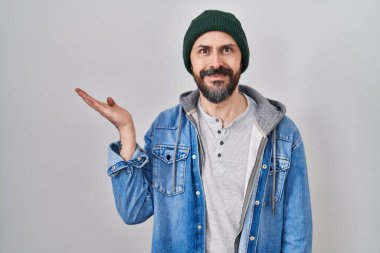 Young hispanic man with tattoos wearing wool cap smiling cheerful presenting and pointing with palm of hand looking at the camera. 