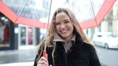 Young beautiful hispanic woman smiling confident holding umbrella at street