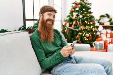 Genç kızıl saçlı adam akıllı telefon kullanıyor. Yılbaşı ağacının yanında oturuyor.