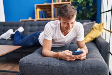 Evdeki kanepede uzanmış akıllı telefon kullanan genç adam.