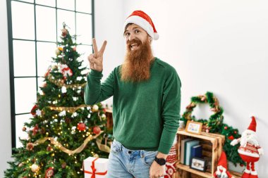 Kızıl saçlı, uzun sakallı, Noel ağacının yanında Noel şapkası takan, kameraya bakıp zafer işareti yapan bir adam. İki numara.. 