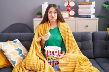 Young hispanic woman eating snack sitting on the sofa at home puffing cheeks with funny face. mouth inflated with air, catching air. 