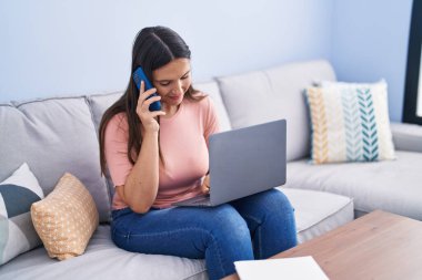 Genç, güzel, İspanyol bir kadın akıllı telefonuyla evde dizüstü bilgisayar kullanıyor.