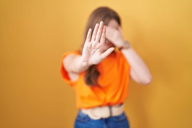 Caucasian woman standing over yellow background covering eyes with hands and doing stop gesture with sad and fear expression. embarrassed and negative concept. 