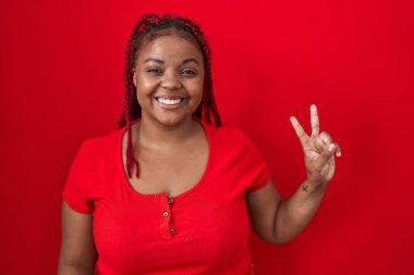 African american woman with braided hair standing over red background smiling with happy face winking at the camera doing victory sign. number two. 