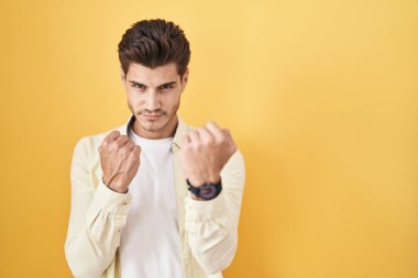 Young hispanic man standing over yellow background ready to fight with fist defense gesture, angry and upset face, afraid of problem 