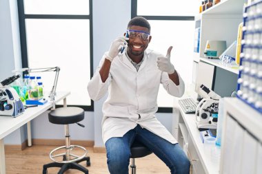 African american man working at scientist laboratory speaking on the phone smiling happy and positive, thumb up doing excellent and approval sign 