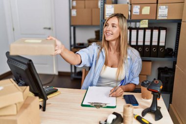 Genç sarışın kadın ecommerce iş işçisi. Ofisteki panoda sipariş paketi yazıyor.