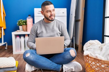 Young latin man using laptop waiting for washing machine at laundry room