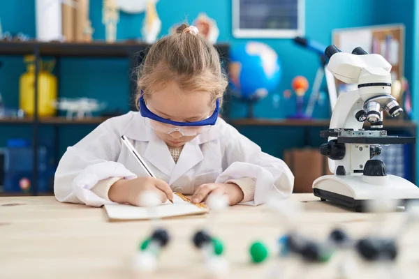 Adorable blonde girl student using microscope writing on notebook at laboratory classroom