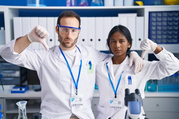 Interracial couple working at scientist laboratory with angry face, negative sign showing dislike with thumbs down, rejection concept