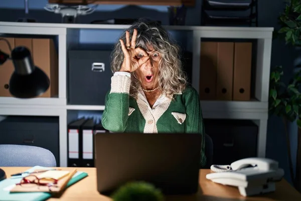 Middle age woman working at night using computer laptop doing ok gesture shocked with surprised face, eye looking through fingers. unbelieving expression.