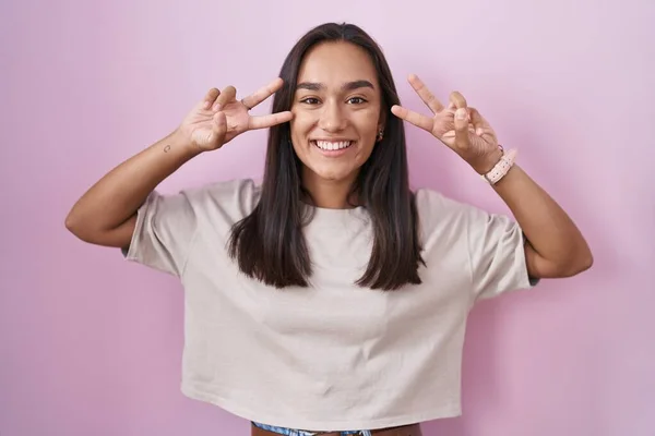 ピンクの背景の上に立つ若いヒスパニック系の女性は 顔の上に指で平和のシンボルを行います 勝利を示す明るい笑顔 — ストック写真
