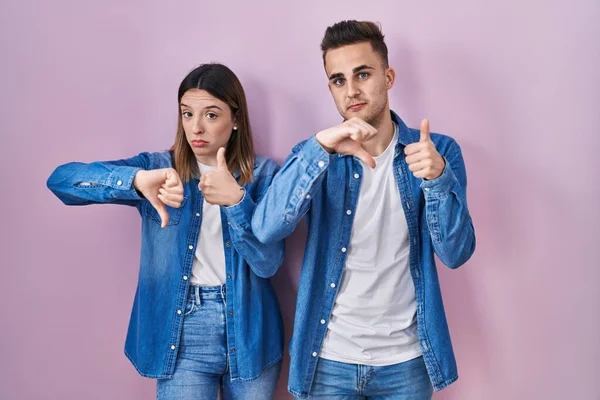 ピンクの背景の上に立つ若いヒスパニック系のカップルは 親指を上下に 意見の相違や合意表現を行う 狂気の葛藤 — ストック写真