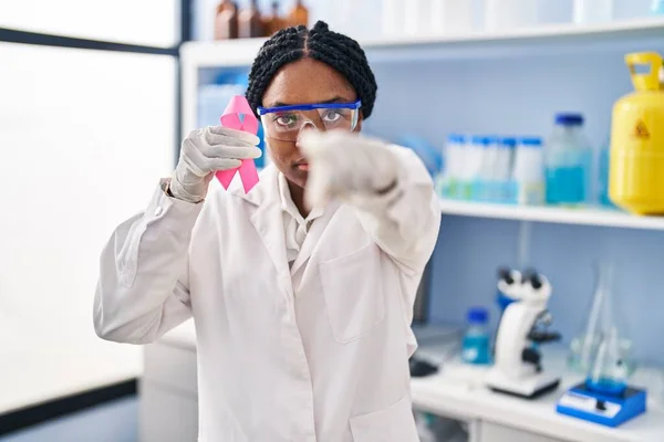 African american woman working at scientist laboratory holding pink ribbon pointing with finger to the camera and to you, confident gesture looking serious