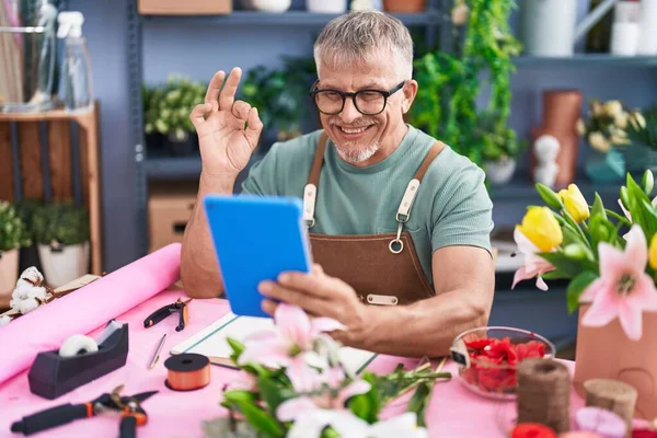 Hispanic man with grey hair working at florist shop doing video call with tablet doing ok sign with fingers, smiling friendly gesturing excellent symbol