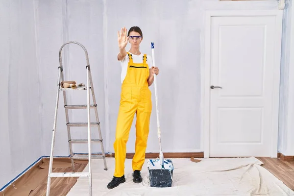 Young caucasian woman painting walls doing stop sing with palm of the hand. warning expression with negative and serious gesture on the face.