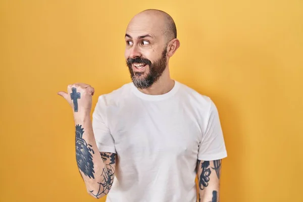 Young hispanic man with tattoos standing over yellow background smiling with happy face looking and pointing to the side with thumb up.