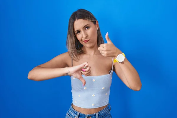 Young woman standing over blue background doing thumbs up and down, disagreement and agreement expression. crazy conflict