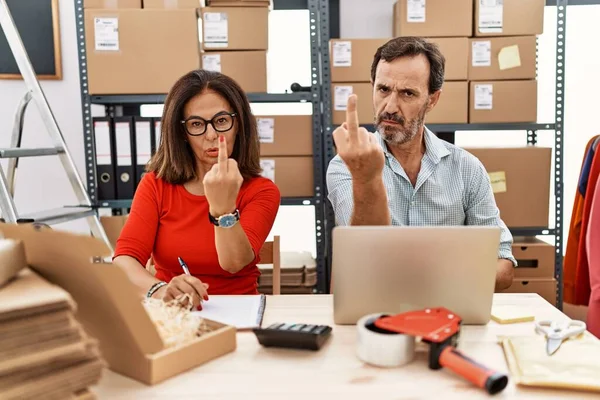 Middle age couple working at small business ecommerce showing middle finger, impolite and rude fuck off expression