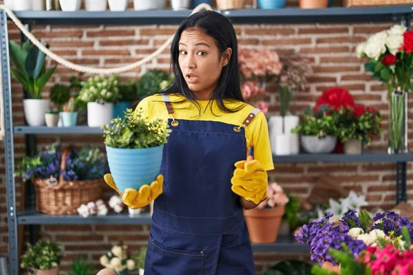 Hispanic woman working at florist shop holding plant in shock face, looking skeptical and sarcastic, surprised with open mouth