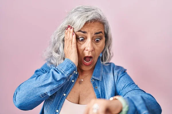 Middle age woman with grey hair standing over pink background looking at the watch time worried, afraid of getting late