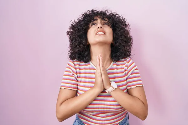 Young middle east woman standing over pink background begging and praying with hands together with hope expression on face very emotional and worried. begging.