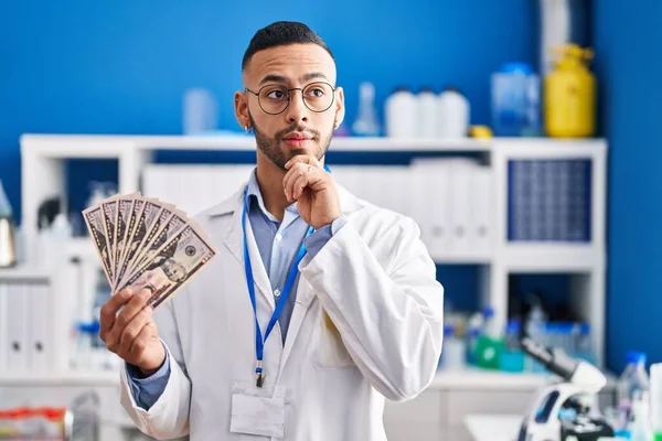 Young Hispanic Man Working Scientist Laboratory Holding Money Serious Face — Stok fotoğraf