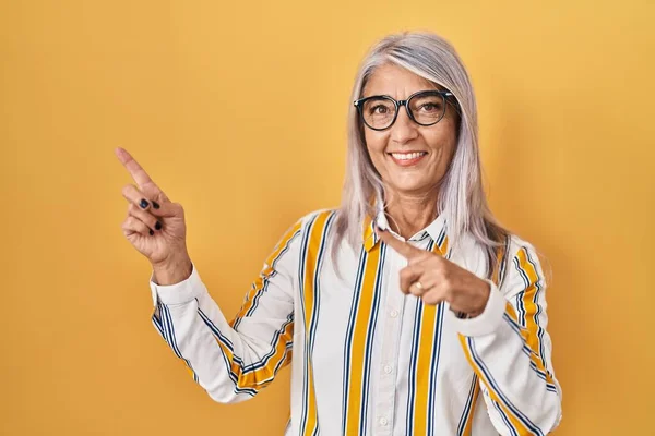 Middle age woman with grey hair standing over yellow background wearing glasses smiling and looking at the camera pointing with two hands and fingers to the side.