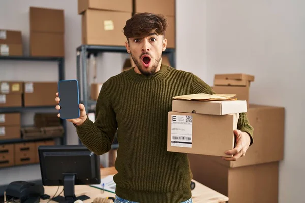 Arab man with beard working at small business ecommerce holding delivery packages in shock face, looking skeptical and sarcastic, surprised with open mouth