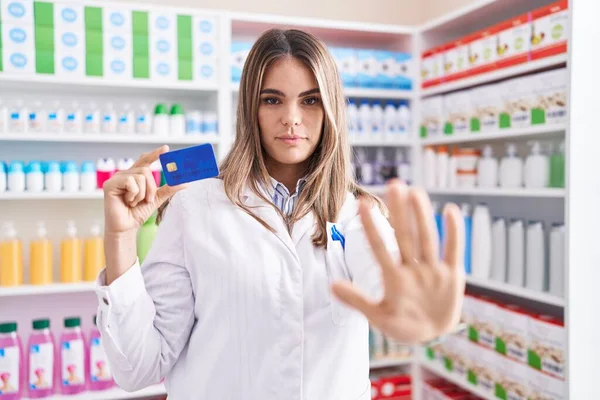 Hispanic young woman working at pharmacy drugstore holding credit card with open hand doing stop sign with serious and confident expression, defense gesture