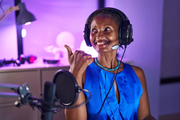 African woman with dreadlocks wearing headphones pointing thumb up to the side smiling happy with open mouth