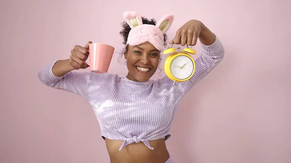戴着睡眠面具的非洲裔美国妇女在孤立的粉色背景下拿着一杯咖啡和闹钟 — 图库照片