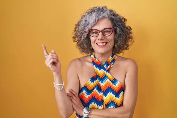 Middle age woman with grey hair standing over yellow background with a big smile on face, pointing with hand finger to the side looking at the camera.
