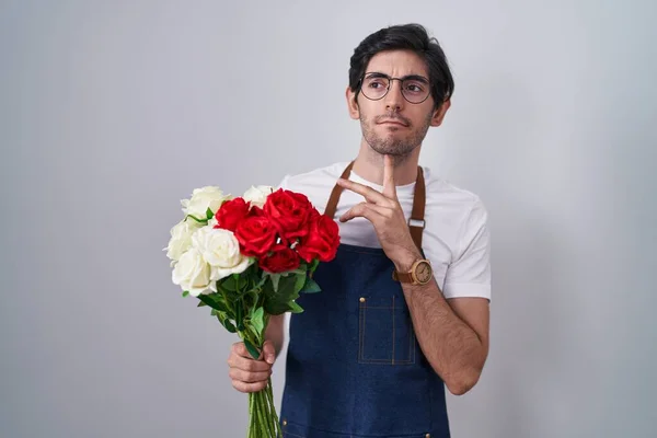 Young hispanic man holding bouquet of white and red roses thinking concentrated about doubt with finger on chin and looking up wondering