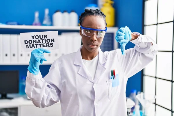Beautiful black woman working at scientist laboratory holding your donation matters banner with angry face, negative sign showing dislike with thumbs down, rejection concept