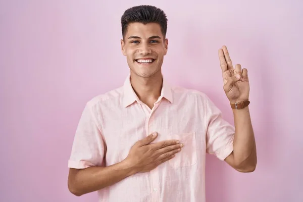 ピンクの背景の上に立つ若いヒスパニック系の男は 胸と指を手で身に着けて笑って 忠誠の誓いをする — ストック写真
