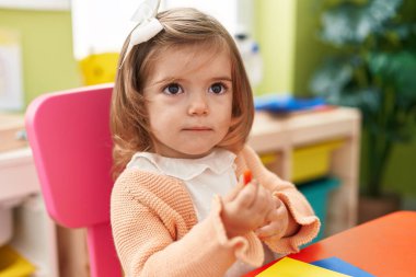 Sevimli sarışın anaokulu öğrencisi anaokulunda masaya oturmuş kağıda resim çiziyor.