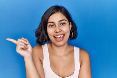 Young hispanic woman standing over blue background with a big smile on face, pointing with hand and finger to the side looking at the camera. 